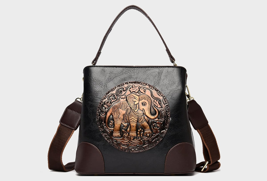 A structured faux-leather handbag featuring a large, central circular bronze plate embossed with an intricate royal Indian design, finished with a slim top handle and a thick brown adjustable strap.