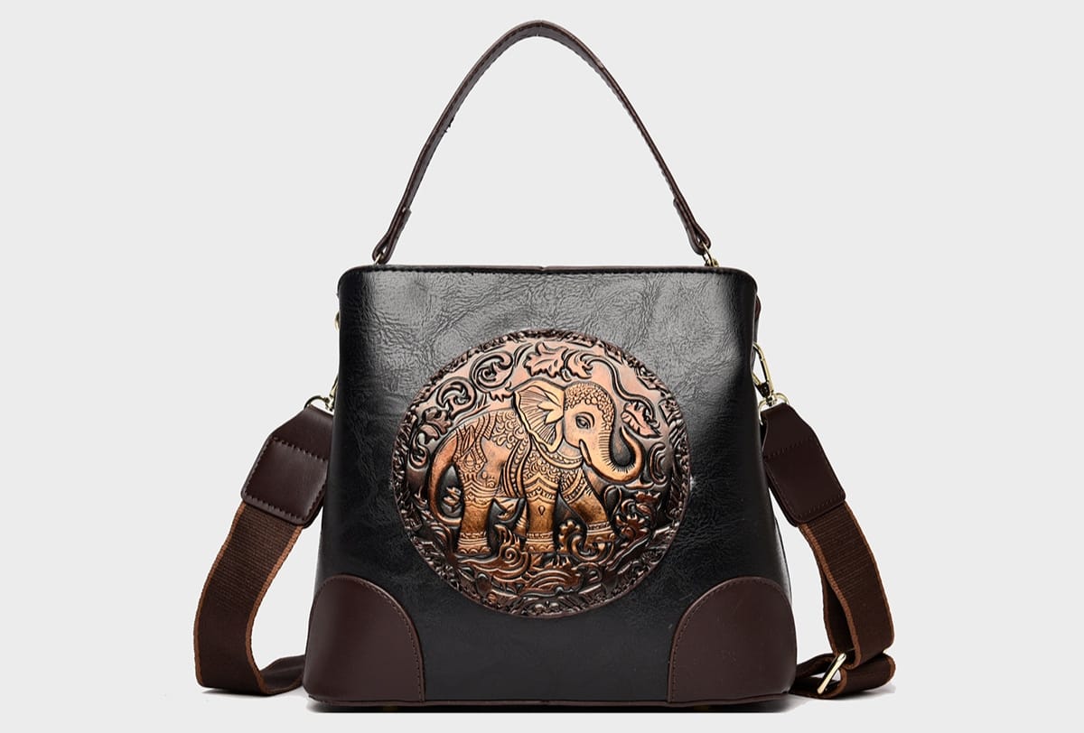 A structured faux-leather handbag featuring a large, central circular bronze plate embossed with an intricate royal Indian design, finished with a slim top handle and a thick brown adjustable strap.