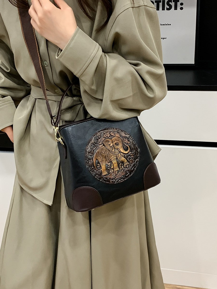 A structured faux-leather handbag featuring a large, central circular bronze plate embossed with an intricate royal Indian design, finished with a slim top handle and a thick brown adjustable strap.