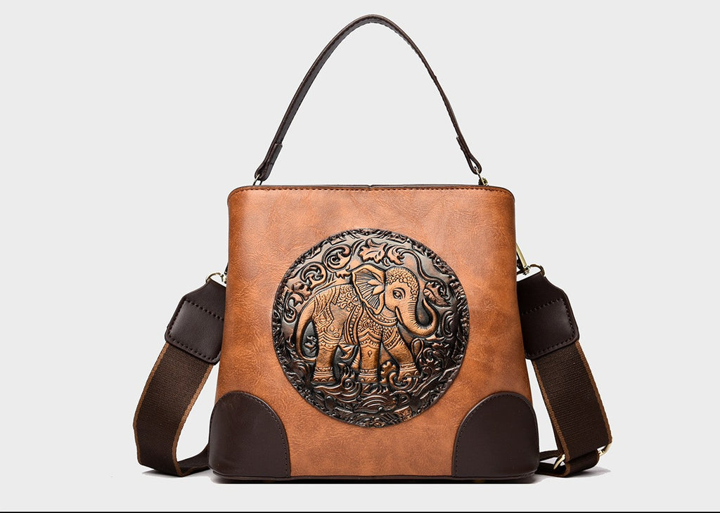 A structured faux-leather handbag featuring a large, central circular bronze plate embossed with an intricate royal Indian design, finished with a slim top handle and a thick brown adjustable strap.