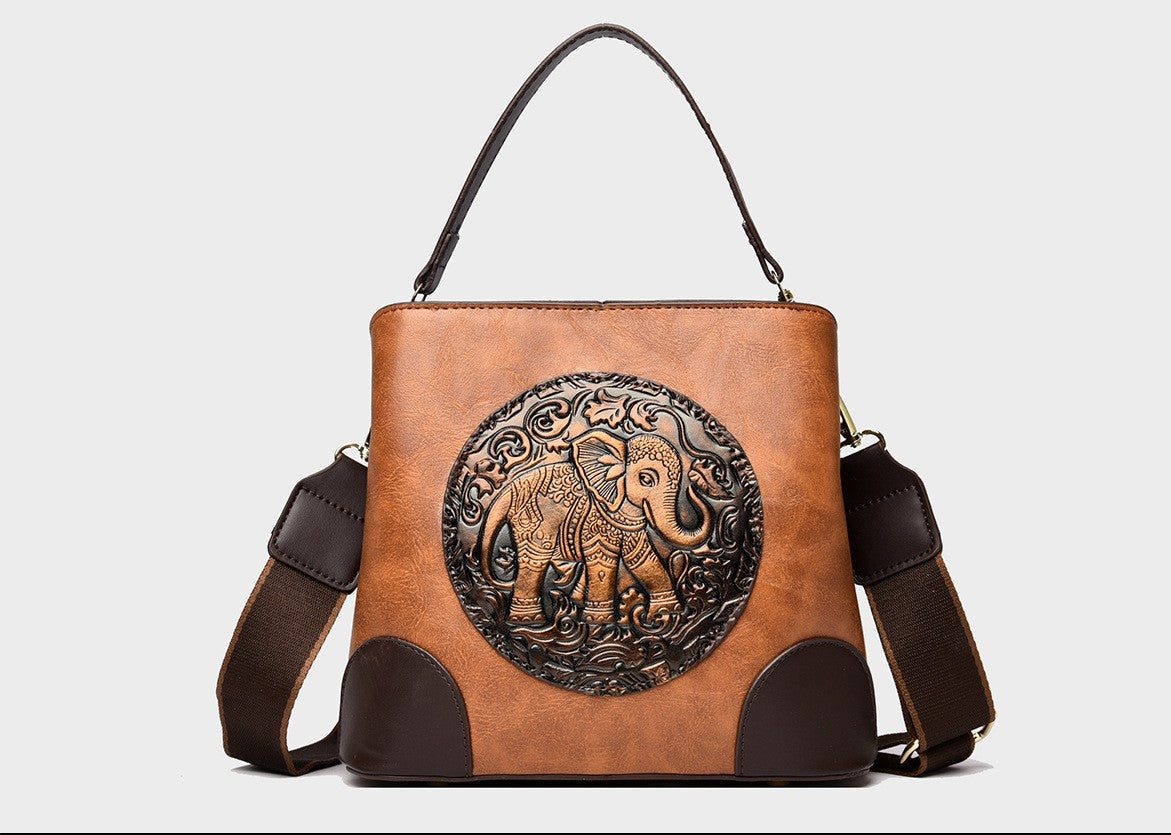 A structured faux-leather handbag featuring a large, central circular bronze plate embossed with an intricate royal Indian design, finished with a slim top handle and a thick brown adjustable strap.