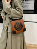 A structured faux-leather handbag featuring a large, central circular bronze plate embossed with an intricate royal Indian design, finished with a slim top handle and a thick brown adjustable strap.