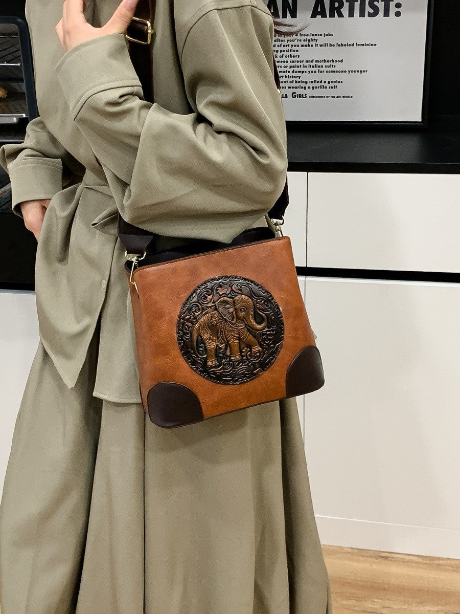 A structured faux-leather handbag featuring a large, central circular bronze plate embossed with an intricate royal Indian design, finished with a slim top handle and a thick brown adjustable strap.