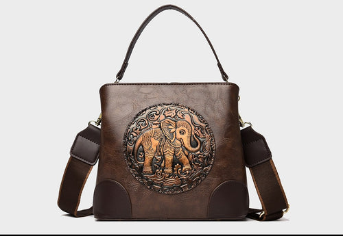 A structured faux-leather handbag featuring a large, central circular bronze plate embossed with an intricate royal Indian design, finished with a slim top handle and a thick brown adjustable strap.