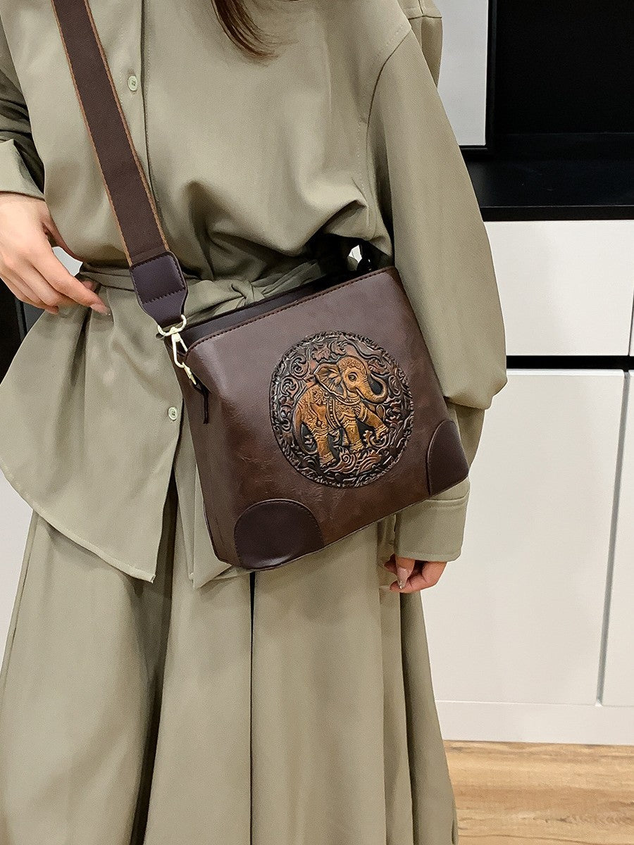 A structured faux-leather handbag featuring a large, central circular bronze plate embossed with an intricate royal Indian design, finished with a slim top handle and a thick brown adjustable strap.