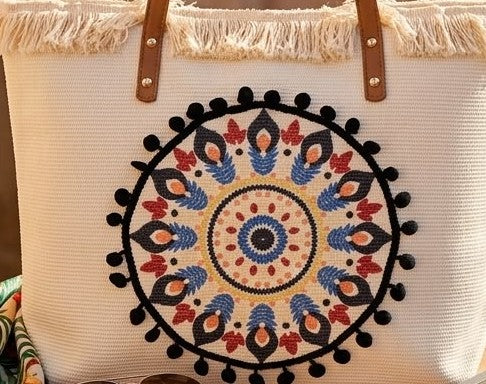 A large canvas tote bag featuring an embroidered geometric mandala circular design on the front panel with a fringed trim along the top rim and leather shoulder handles.