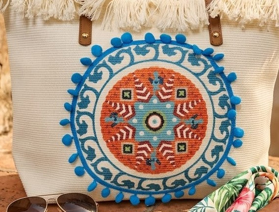A large canvas tote bag featuring an embroidered geometric mandala circular design on the front panel with a fringed trim along the top rim and leather shoulder handles.