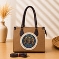 A structured rectangular canvas satchel handbag featuring an intricate circular fringed emblem with an artisanal motif, accented by contrast top handles and a matching detachable shoulder strap.