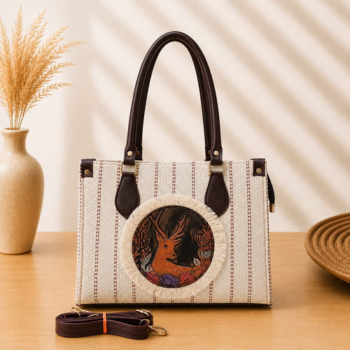 A structured rectangular canvas satchel handbag featuring an intricate circular fringed emblem with an artisanal motif, accented by contrast top handles and a matching detachable shoulder strap.